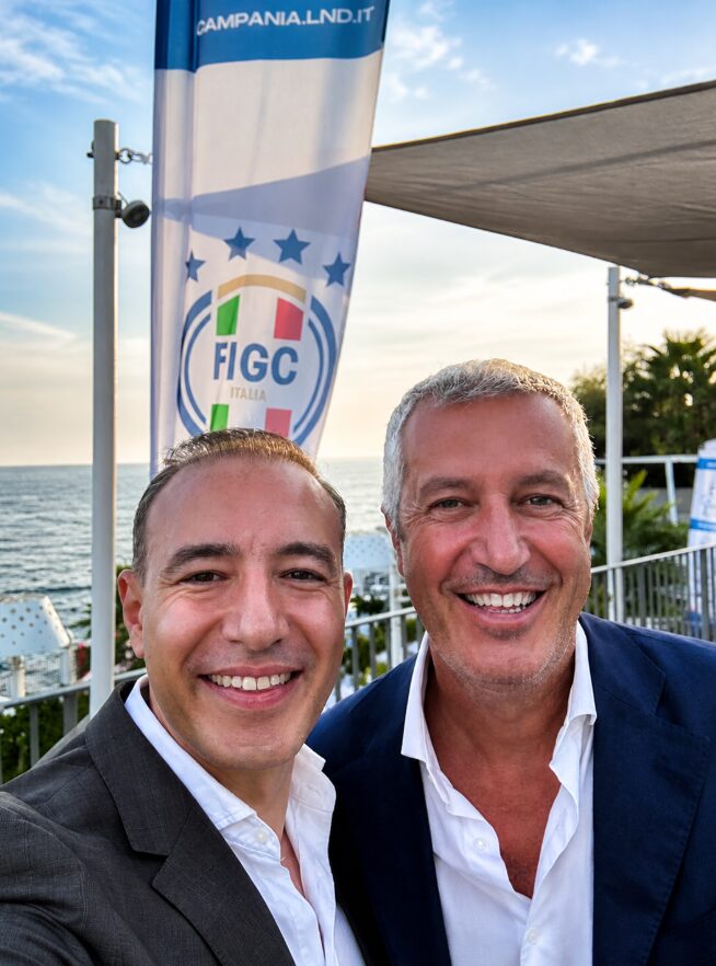 Marco Cioffi con un dirigente del calcio italiano davanti alla bandiera della FIGC — Lega Nazionale Dilettanti Campania, in un evento sul mare.
