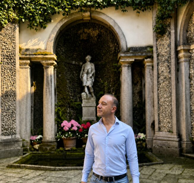 Marco Cioffi nel ninfeo barocco dei giardini dell'Isola Bella sul Lago Maggiore, davanti a una statua incorniciata da un'arcata in pietra e ortensie.