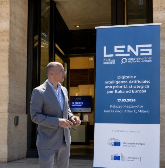 Marco Cioffi davanti all'ingresso di Palazzo Mezzanotte a Milano, accanto al totem dell'evento LENS 2026 degli Osservatori Digital Innovation del Politecnico di Milano.