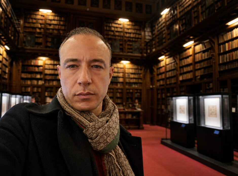 Marco Cioffi nella sala storica della Biblioteca Ambrosiana di Milano, circondato da scaffali di volumi antichi e teche con manoscritti.